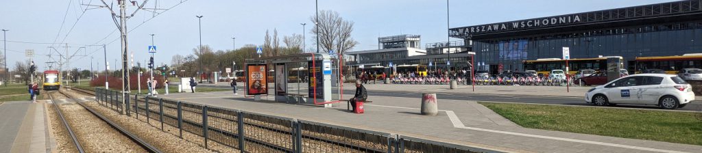 Widok na dworzec Warszawa Wschodnia z przystanku tramwajowego. W tle widoczni przechodnie, nadjeżdżający tramwaj, autobusy oraz rowery miejskie